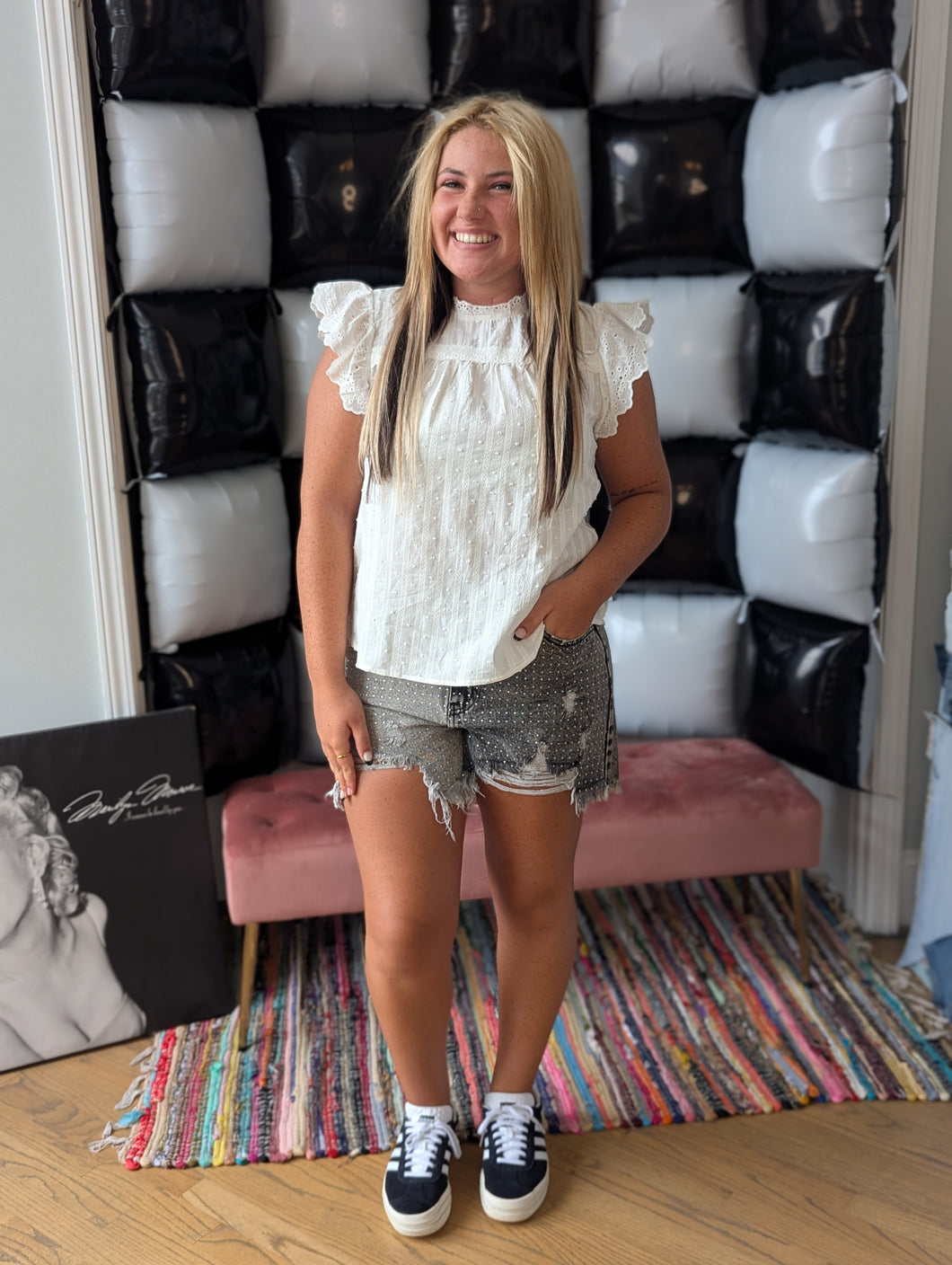 A woman standing in front of a black and white decorative wall, wearing a white eyelet top with ruffle sleeves and denim shorts, with sneakers on her feet.